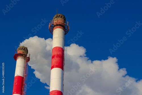 Red and white pipe of boiler plant with white steam and pipe without smoke against blue sky. Heating season in winter. Industrial background. Air pollution from fuel combustion