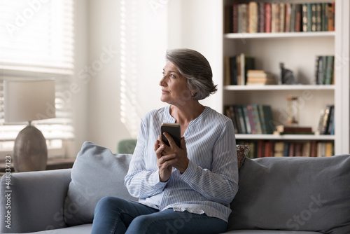 Dreamy middle aged woman looking in distance, holding smartphone in hands, thinking of getting positive message, feeling relaxed using cellphone software application sitting on sofa alone at home.
