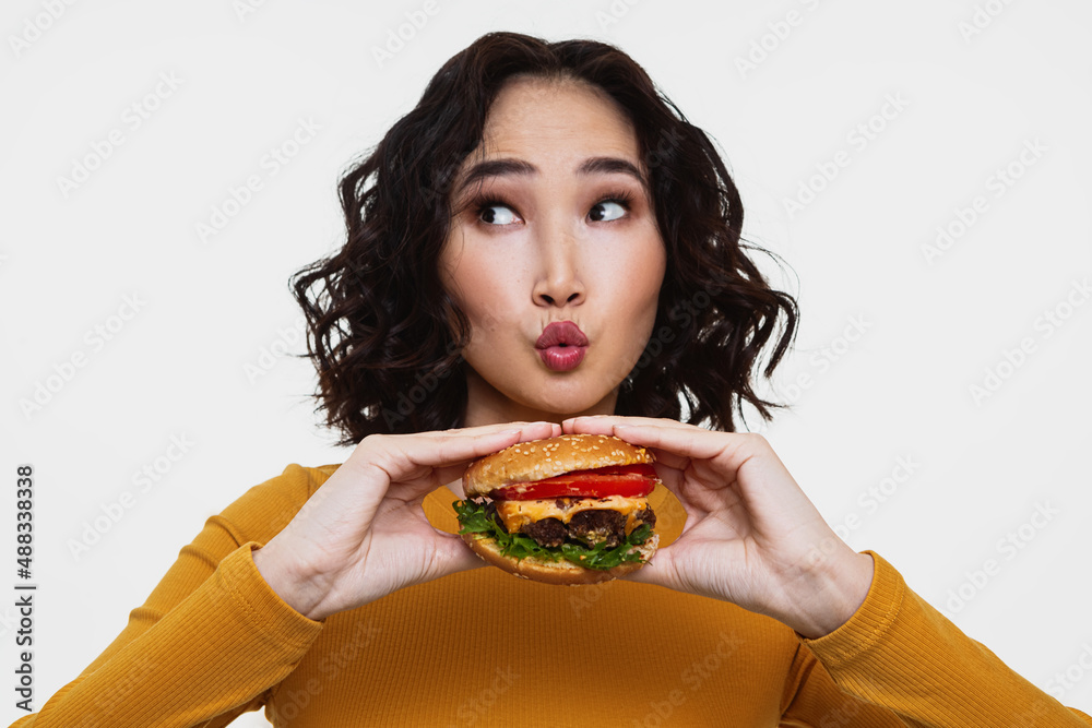 A horizontal photo of a pretty curly Asian girl protrude her lips looking away, she’s holding a cheeseburger with two hands A horizontal photo of a pretty curly Asian girl protrude her lips looking away, she’s holding a cheeseburger with two hands
