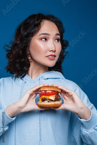 A young curly smiling Asian woman with bright makeup looking away wearing a blue hoodie holding a big homemade burger with two hands over a navy blue background