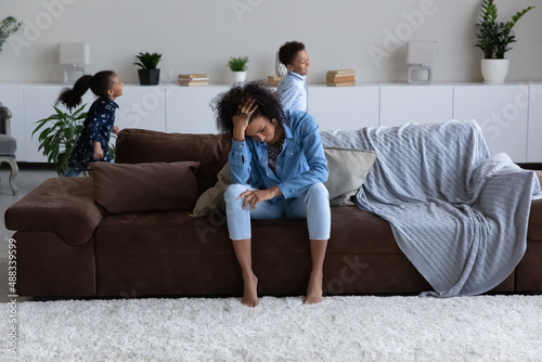 Frustrated millennial African American mum or babysitter touching forehead, suffering from headache, feeling tired of noisy small kids boy girl having fun in modern living room, generations gap.