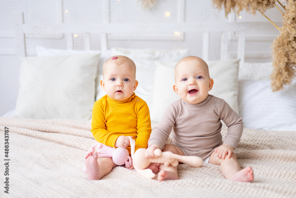 sister babies are sitting on the bed at home and smiling while playing, two cute little twin babies in a cotton bodysuit with hemangioma sister babies are sitting on the bed at home and smiling while playing, two cute little twin babies in a cotton bodysuit with hemangioma