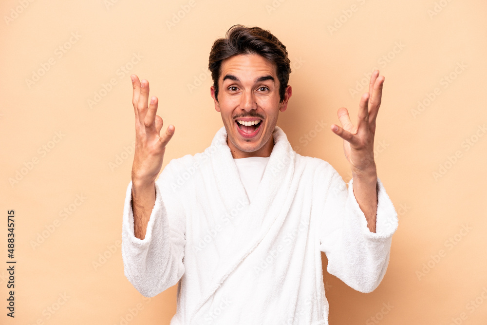 Young caucasian man wearing a bathrobe isolated on beige background receiving a pleasant surprise, excited and raising hands. Young caucasian man wearing a bathrobe isolated on beige background receiving a pleasant surprise, excited and raising hands.