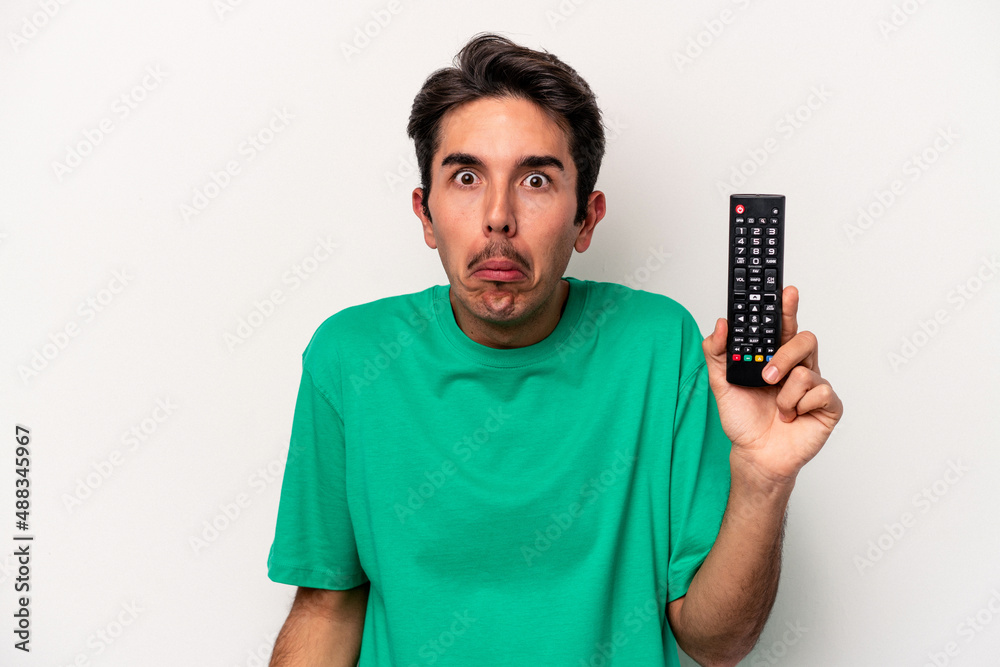 Young caucasian man holding a tv remote isolated on white background shrugs shoulders and open eyes confused. Young caucasian man holding a tv remote isolated on white background shrugs shoulders and open eyes confused.