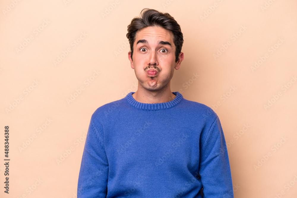 Young caucasian man isolated on beige background blows cheeks, has tired expression. Facial expression concept. Young caucasian man isolated on beige background blows cheeks, has tired expression. Facial expression concept.