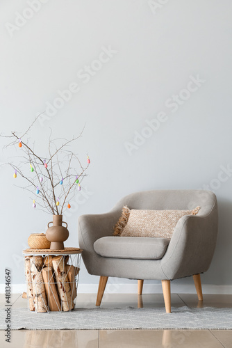 Tree branches with colorful Easter eggs on table and armchair near light wall photo