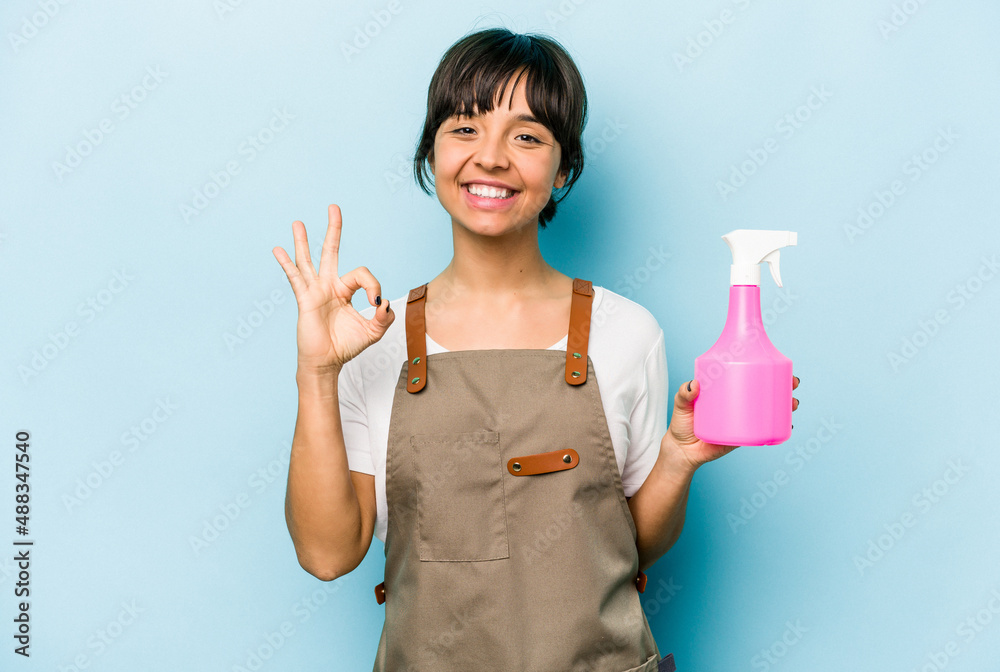 Young hispanic gardener woman holding a hand sprayer isolated on blue background cheerful and confident showing ok gesture. Young hispanic gardener woman holding a hand sprayer isolated on blue background cheerful and confident showing ok gesture.