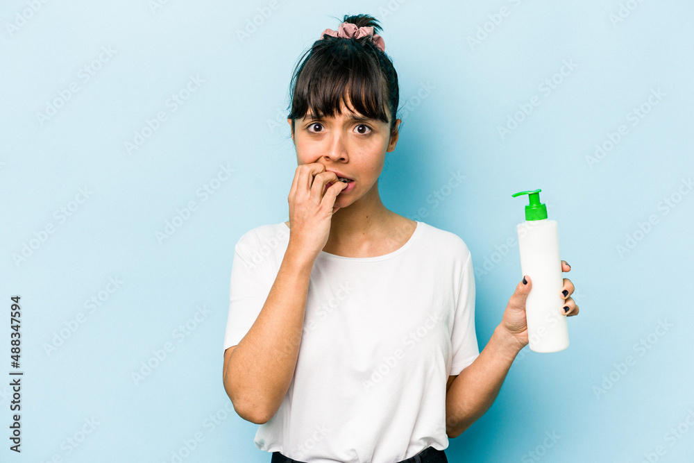 Young hispanic woman holding a body lotion isolated on blue background biting fingernails, nervous and very anxious. Young hispanic woman holding a body lotion isolated on blue background biting fingernails, nervous and very anxious.