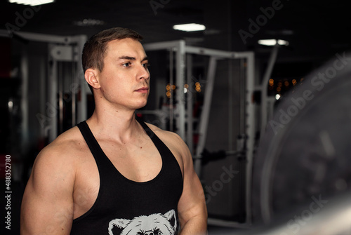 Close up portrait of athletic muscled young man in black t-shirt standing among sport gym stuff on black grey dark background