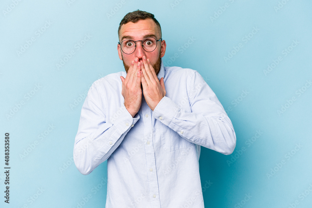 Young caucasian man isolated on blue background shocked covering mouth with hands. Young caucasian man isolated on blue background shocked covering mouth with hands.