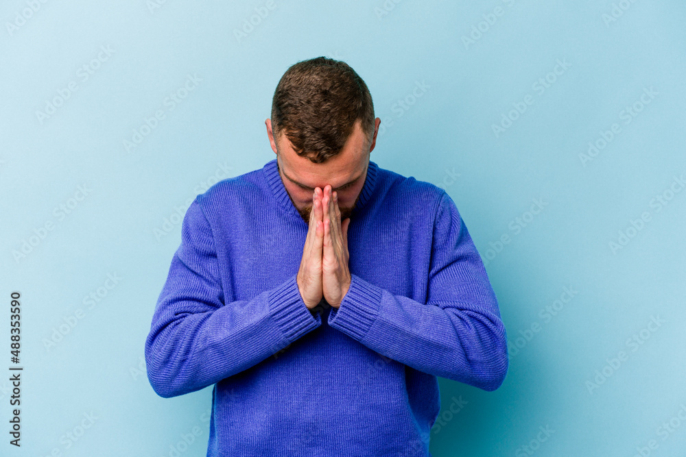Young caucasian man isolated on blue background praying, showing devotion, religious person looking for divine inspiration. Young caucasian man isolated on blue background praying, showing devotion, religious person looking for divine inspiration.