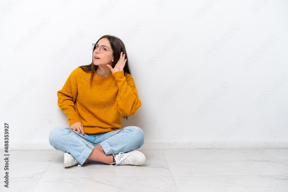 Young girl sitting on the floor isolated on white background listening to something by putting hand on the ear