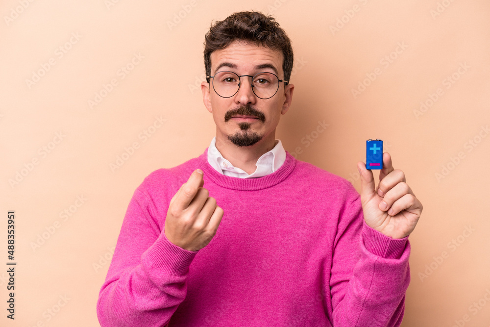 Young caucasian man holding batteries isolated on beige background pointing with finger at you as if inviting come closer. Young caucasian man holding batteries isolated on beige background pointing with finger at you as if inviting come closer.