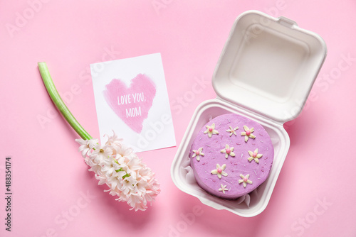 Plastic lunch box with tasty bento cake, greeting card and flowers on pink background