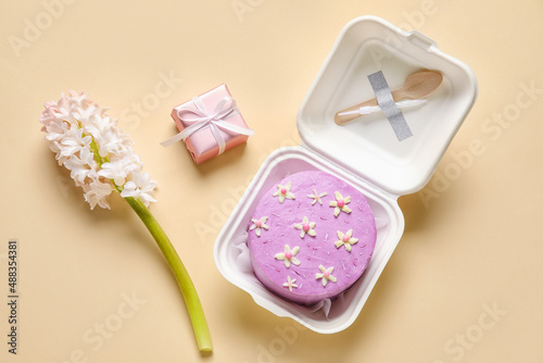Plastic lunch box with tasty bento cake, gift and flowers on beige background