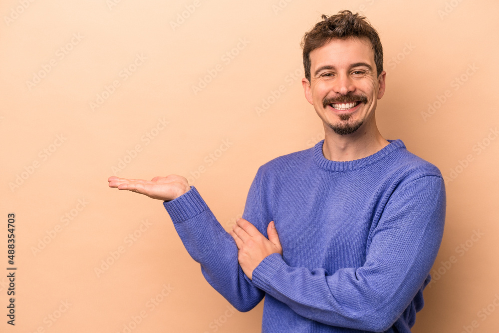 Young caucasian man isolated on beige background showing a copy space on a palm and holding another hand on waist.