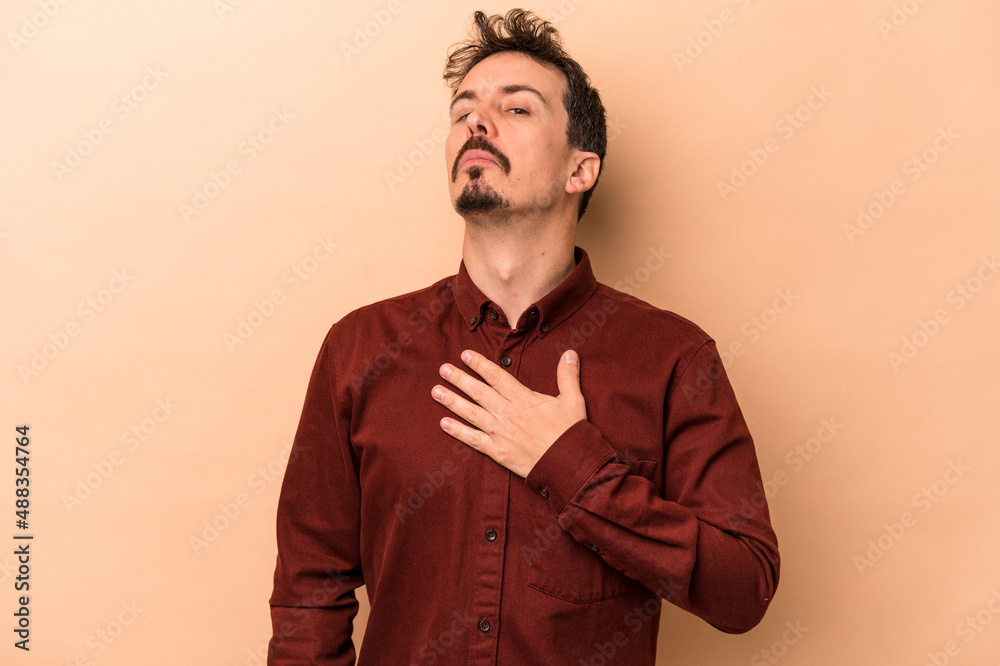 Young caucasian man isolated on beige background taking an oath, putting hand on chest.