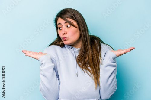Young caucasian overweight woman isolated on blue background confused and doubtful shrugging shoulders to hold a copy space.