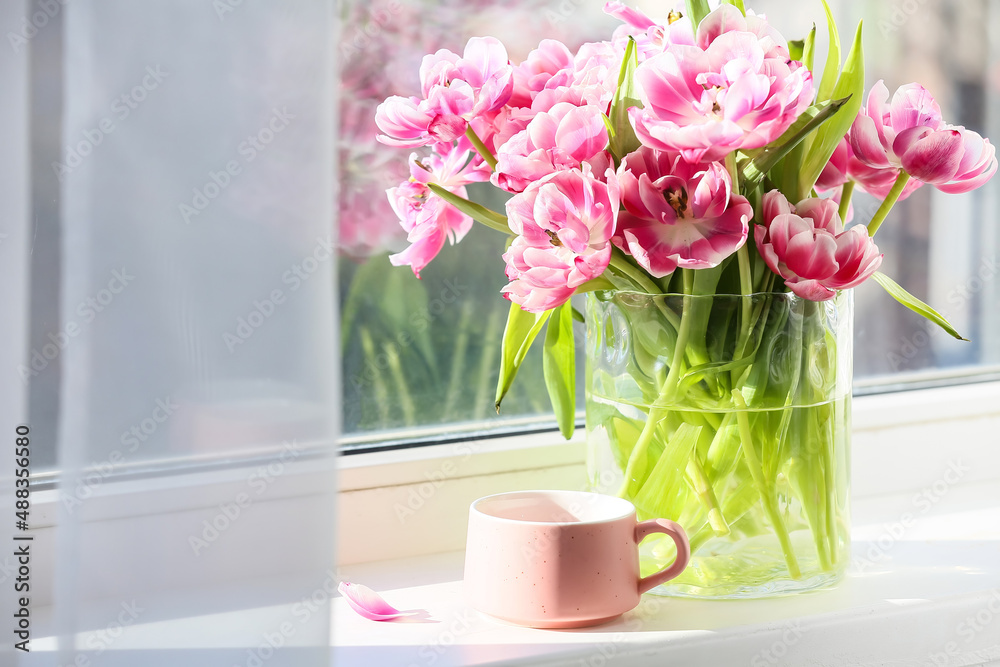 Glass vase with beautiful tulip flowers and cup of tea on windowsill Glass vase with beautiful tulip flowers and cup of tea on windowsill