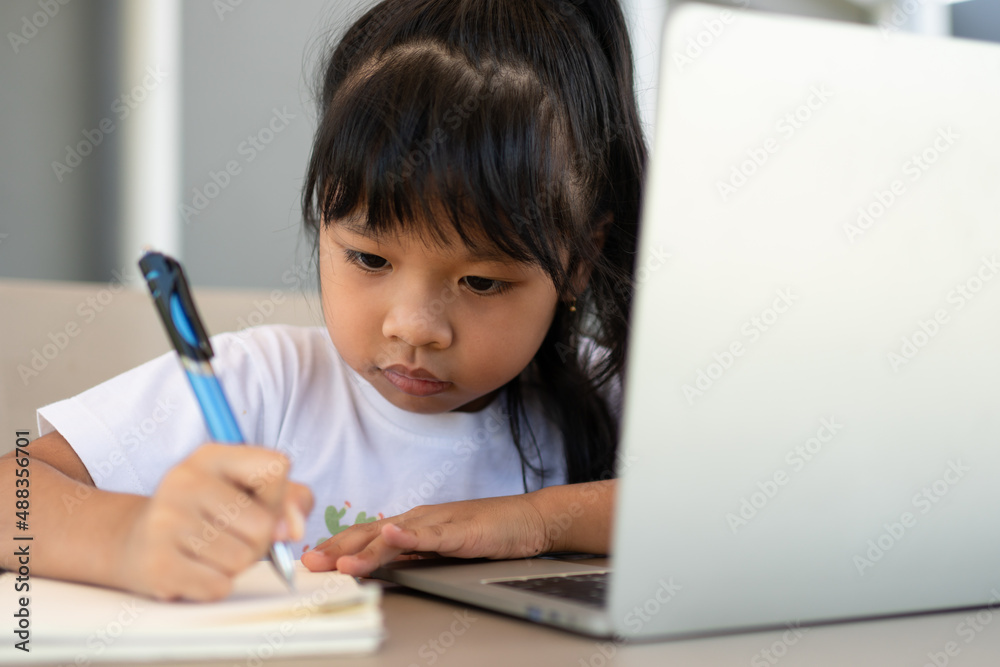 Asian girl student holding a pen for taking notes and doing homework and using the laptop for online learning class at home, Education and distance learning for children Homeschooling concept Asian girl student holding a pen for taking notes and doing homework and using the laptop for online learning class at home, Education and distance learning for children Homeschooling concept
