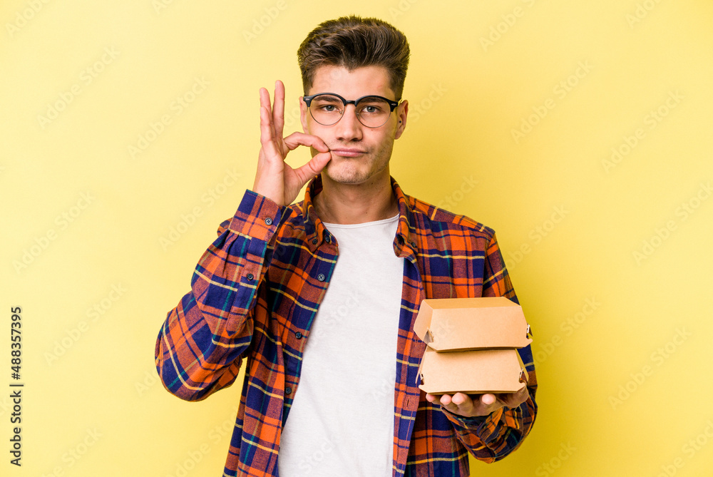 Young caucasian man holding a burger isolated on yellow background with fingers on lips keeping a secret. Young caucasian man holding a burger isolated on yellow background with fingers on lips keeping a secret.