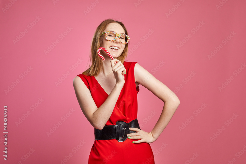 Beautiful sexy young girl in red dress and sweet sprinkles glasses is holding a festive candy cane in hand on a red background. Beautiful sexy young girl in red dress and sweet sprinkles glasses is holding a festive candy cane in hand on a red background.