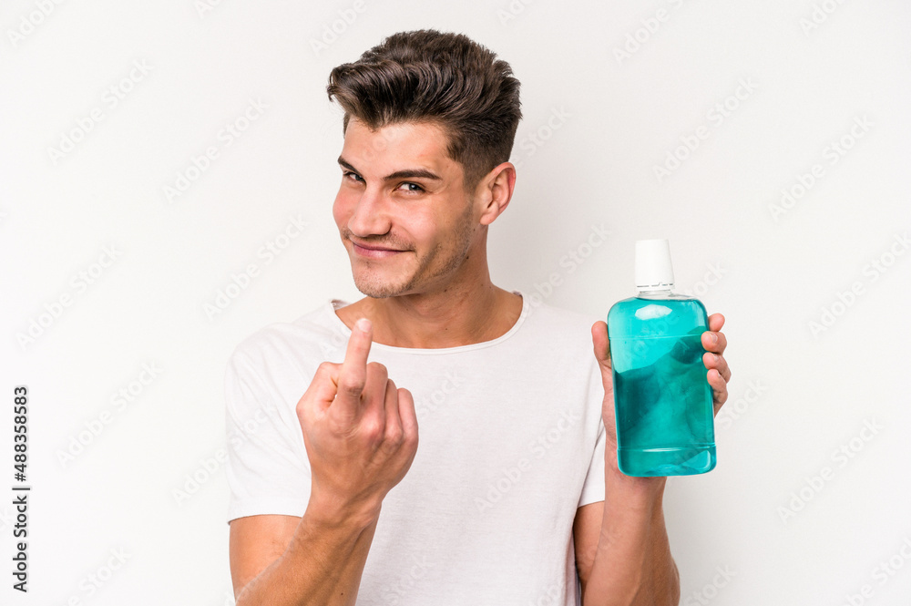 Young caucasian man holding mouthwash isolated on white background pointing with finger at you as if inviting come closer.