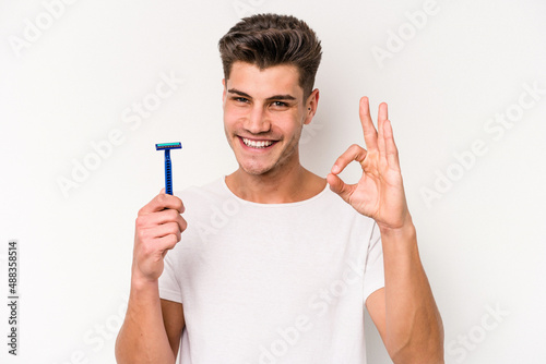 Young caucasian man shaving his beard isolated on white background cheerful and confident showing ok gesture.
