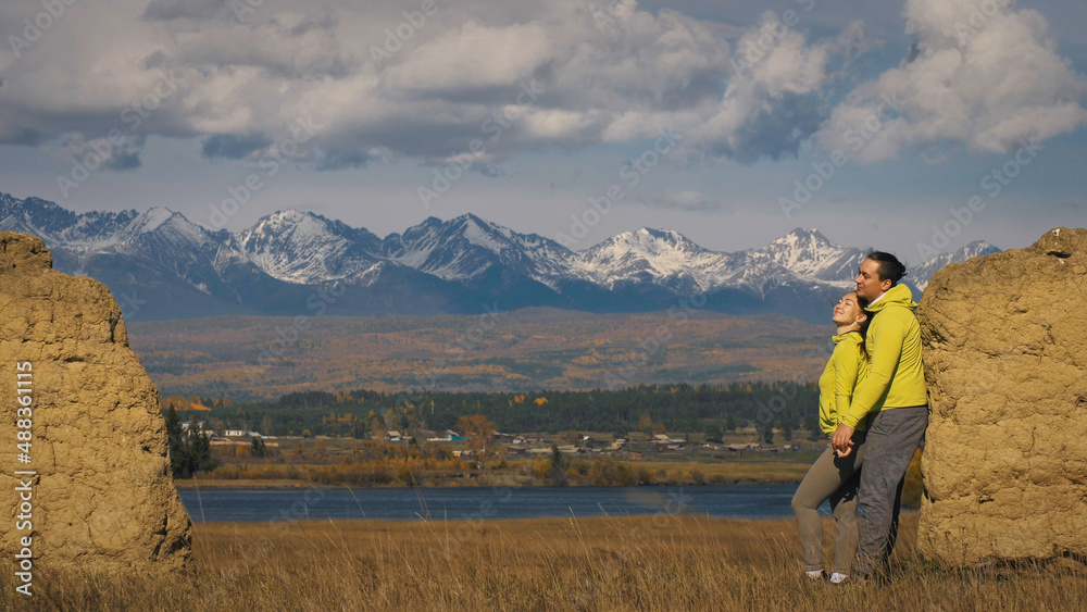 Man and woman in yellow green sportswear. Lovely couple of travelers hug and kiss near old stone enjoying highland landscape. Two travelers are walking against the backdrop of snow-capped mountains. Man and woman in yellow green sportswear. Lovely couple of travelers hug and kiss near old stone enjoying highland landscape. Two travelers are walking against the backdrop of snow-capped mountains.