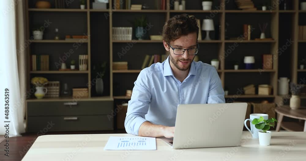 Thoughtful young businessman freelancer in glasses work on statistic report using laptop pc at home office take notes from screen. Busy millennial man worker employee think on stat data analyze sales