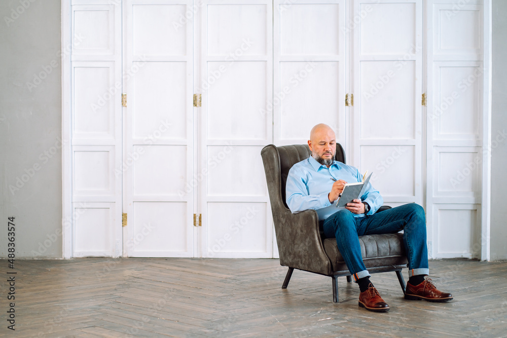 Portrait of confident bald adult man with beard sitting in leather armchair in office, taking notes in datebook planner, thinking new investment. Wearing jeans, shirt, boots. Cabinet closet indoor Portrait of confident bald adult man with beard sitting in leather armchair in office, taking notes in datebook planner, thinking new investment. Wearing jeans, shirt, boots. Cabinet closet indoor