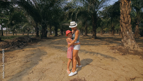 Woman tourist with plait walks looking around at growing young trees with lush leaves at oil palm farm elaeis guineensis on sunny day. Mom and daughter for a walk. Concept of exotic crop cultivation photo