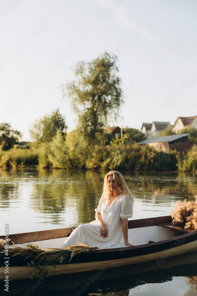 young blonde woman with wavy hair sits in a boat on the lake. girl in a long white dress posing in an old wooden boat. evening boat trip on the water near the house.
