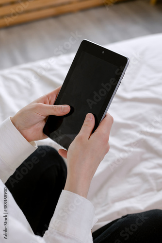 Woman's hands hold a black tablet. Sits on a bed in an apartment.