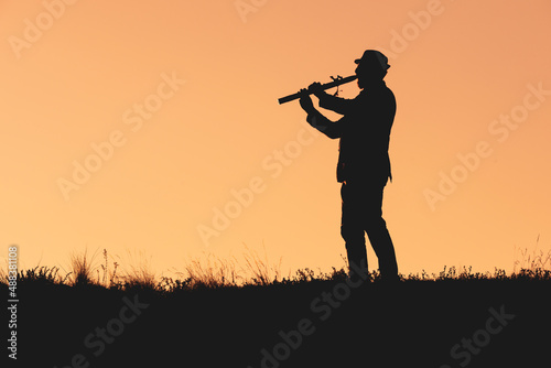 silhouette of a man playing the flute in a hat on an orange background