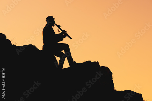 silhouette of a man in a hat sitting on a mountain and playing the flute