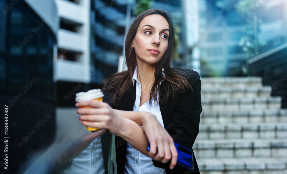 Casually dressed business woman standing on city street and using her smart phone to communicate with someone while holding coffee to go in other hand. Bright sunny day.