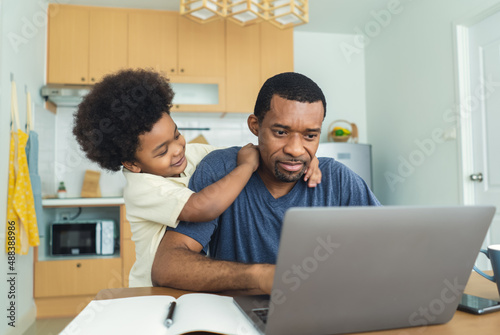 Portriat of busy African American father working from home office using laptop computer sit at kitchen table with cute little son playing nearly to disturb. Exhausted parent with hyperactive child.