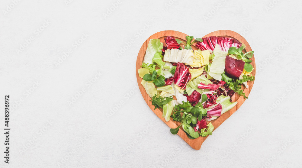 Fototapeta premium Sliced colored lettuce leaves in a wooden plate heart shape on a light cement background.