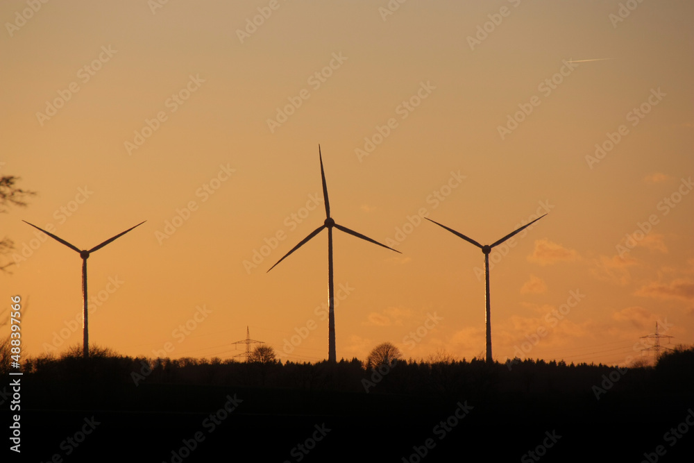Silhouette von Windrädern und Strommasten vor orangenem Sonnenuntergang