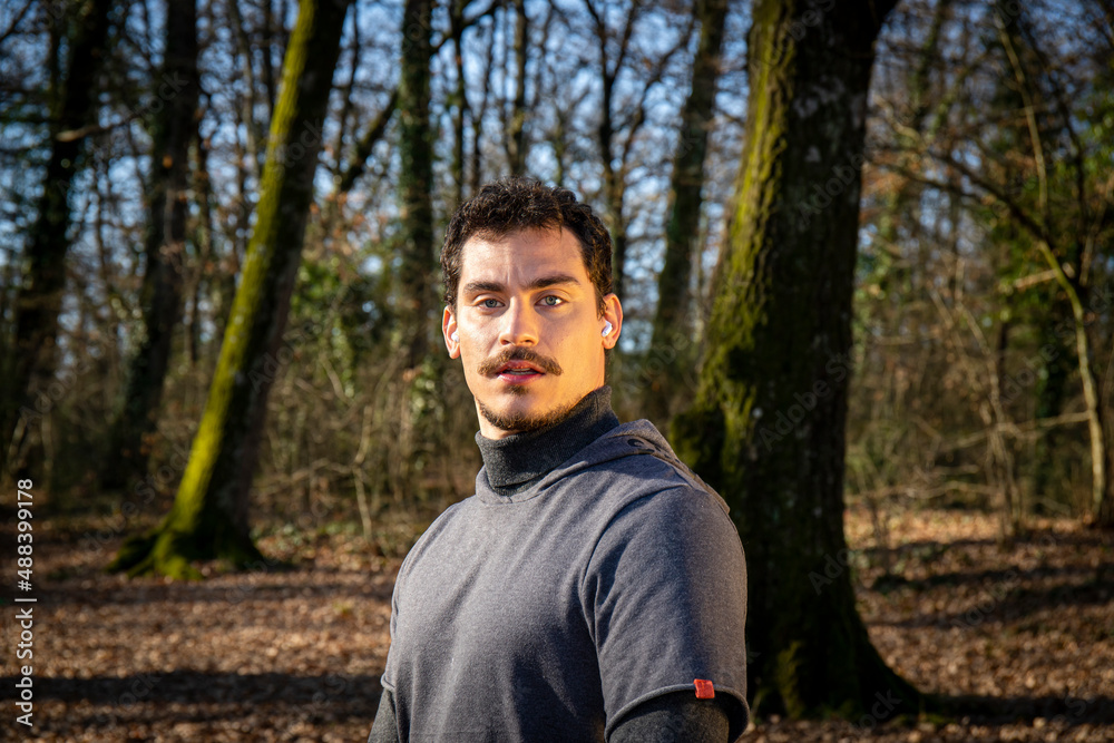 Runner is doing stretching in the woods. Young man with blue eyes and mustache is training in the park.