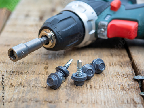 Close-up of a cordless screwdriver lying on a wooden surface with a nozzle for roofing screws. Self-tapping screws painted in gray are lying nearby
