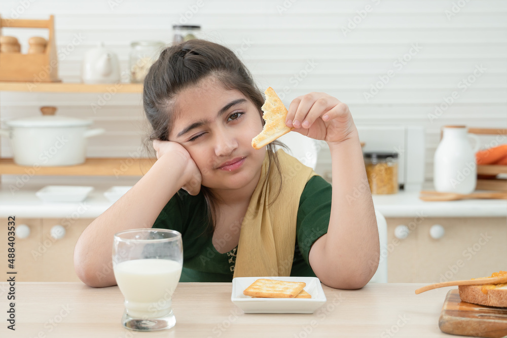 Little cute Indian kid girl holding and eating crackers for breakfast with a glass of milk, jam and bread in kitchen at home. A good diet is important to the growth of children concept Little cute Indian kid girl holding and eating crackers for breakfast with a glass of milk, jam and bread in kitchen at home. A good diet is important to the growth of children concept