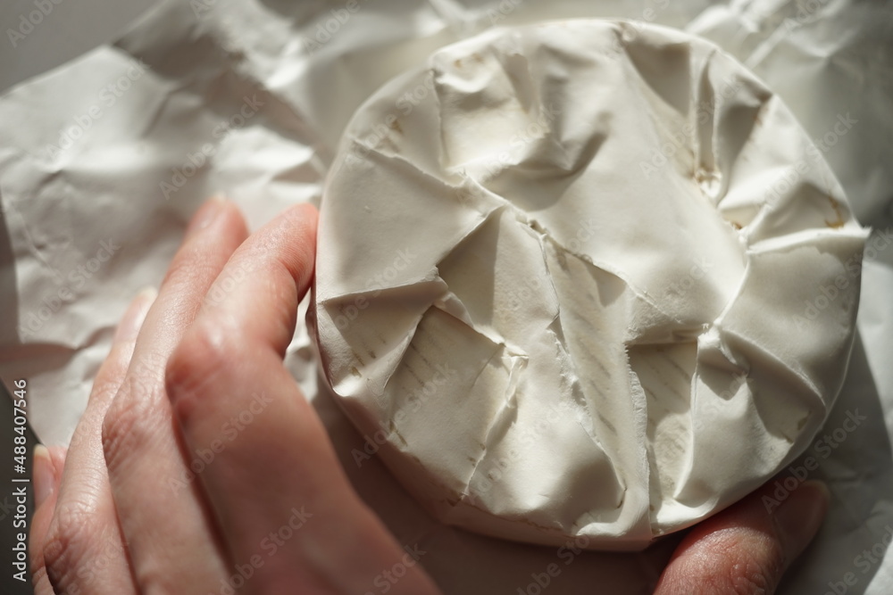Fingers touch whole round camembert cheese on a sunny wrapping paper. Fingers touch whole round camembert cheese on a sunny wrapping paper.