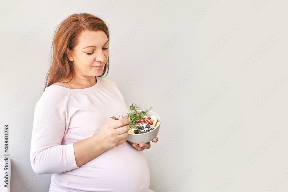 Pregnant woman eating healthy food. Organic calcium salad with fruits and microgreen. Round plate. Home dinner or lunch. Vitamin cereal. Holding bowl in hands. Pregnant woman eating healthy food. Organic calcium salad with fruits and microgreen. Round plate. Home dinner or lunch. Vitamin cereal. Holding bowl in hands.