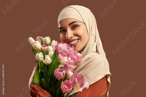 Beautiful Muslim woman in a beige hijab holding a bouquet of white and pink tulips smiling and looking at the camera. Beauty, spring concept. photo