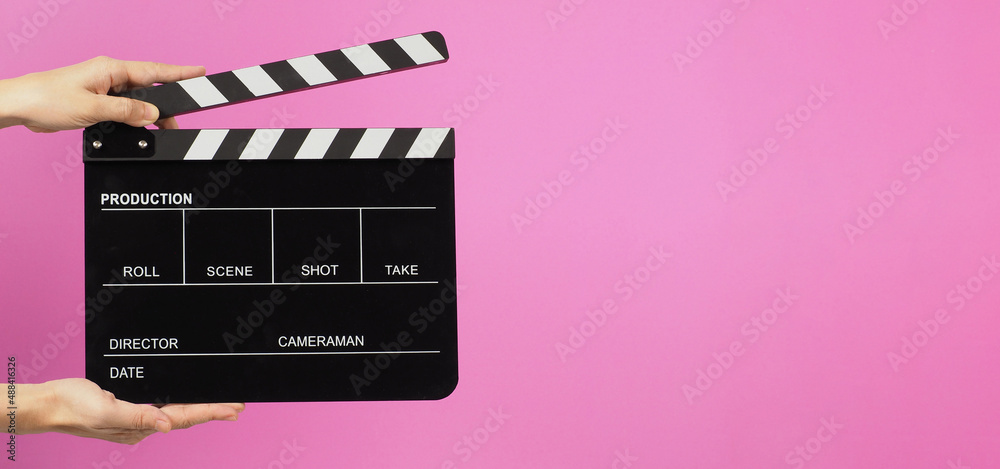 Two hands are holding a black clapper board or movie slate on pink background.