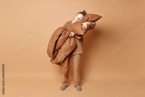 Sleeping routine and wellbeing concept. Relaxed young woman leans on soft pillow wears pajama and slippers blindfold on eyes takes nap isolated over beige background enjoys peaceful atmosphere
