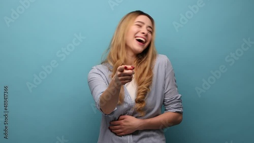 Hey, you are ridiculous. Joyful woman laughing, holding stomach, pointing to camera, taunting you, can't stop hysterical laughter, wearing striped shirt. Indoor studio shot isolated on blue background