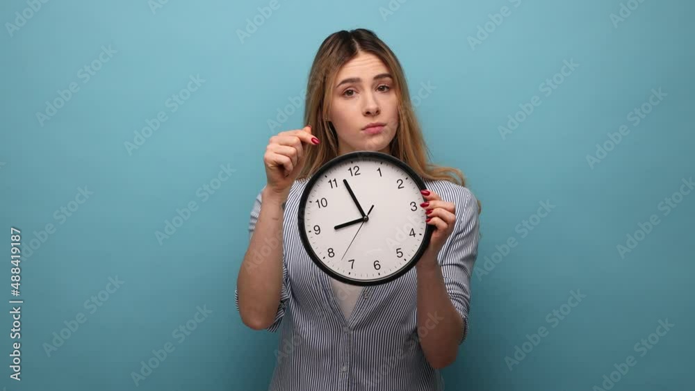 Portrait of serious woman with wavy hair holding big wall clock, looking at camera with unpleasant emotions, time to go, wearing striped shirt. Indoor studio shot isolated on blue background. Portrait of serious woman with wavy hair holding big wall clock, looking at camera with unpleasant emotions, time to go, wearing striped shirt. Indoor studio shot isolated on blue background.